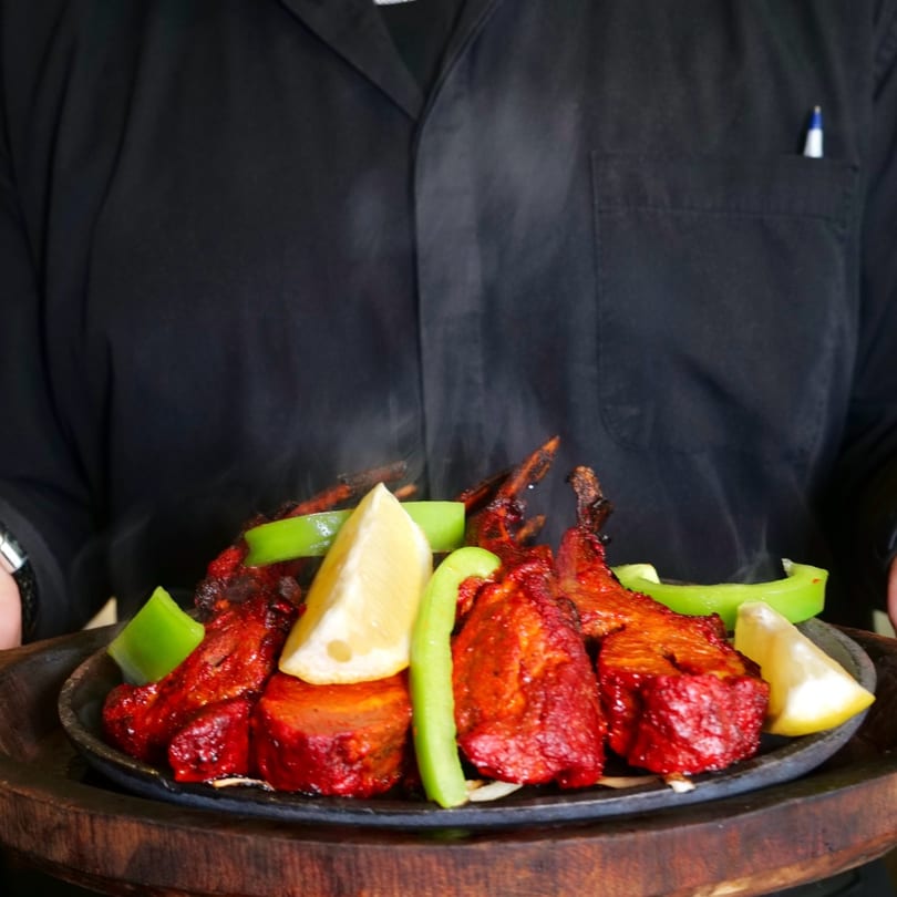 A server holds a sizzling platter of red tandoori chicken with green peppers and lemon wedges.