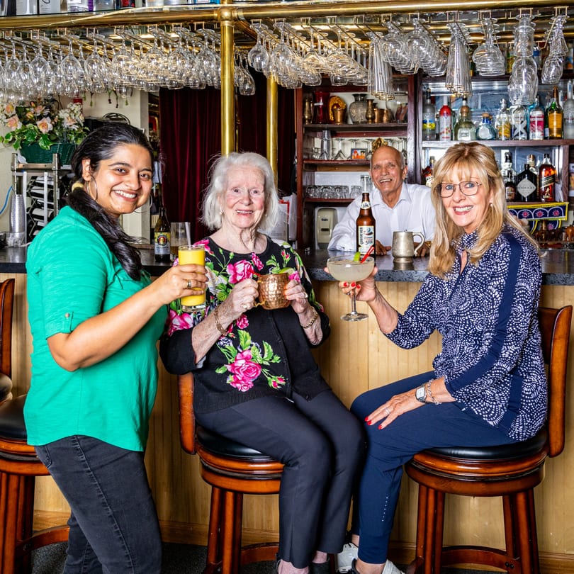 Three women and a man smiling with drinks at the INDIA'S Restaurant bar.