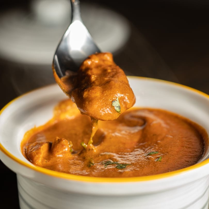 A spoonful of rich, orange curry with herbs being lifted from a white bowl with a yellow rim.
