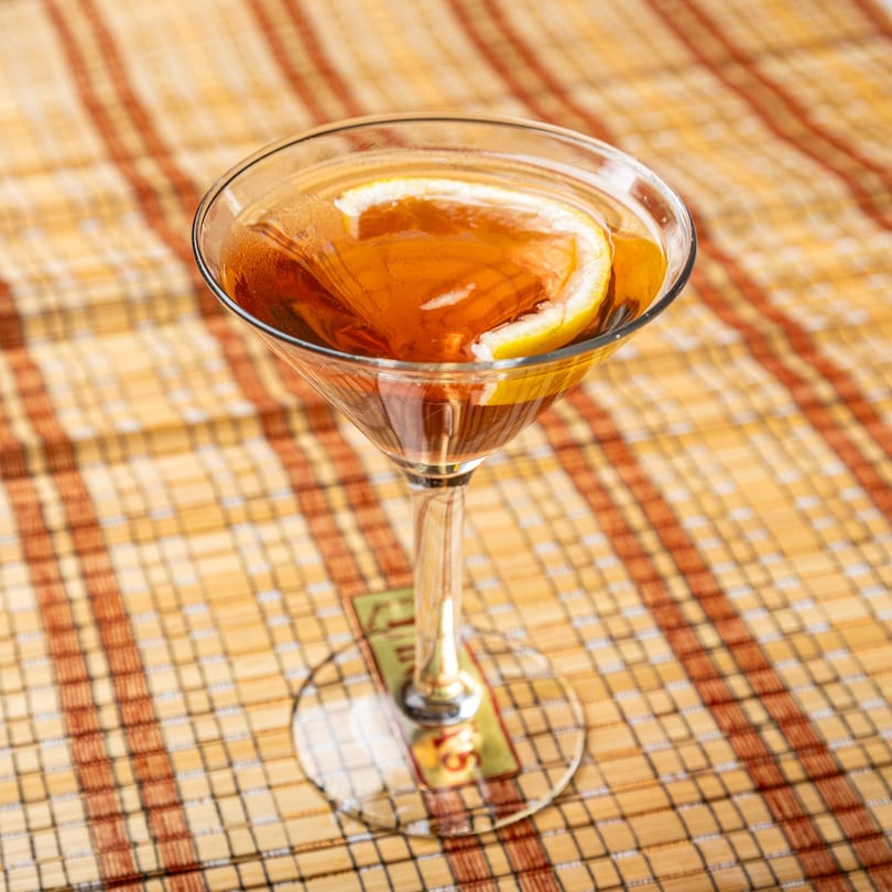 A martini glass filled with a dark amber drink and a lemon slice, on a woven placemat.