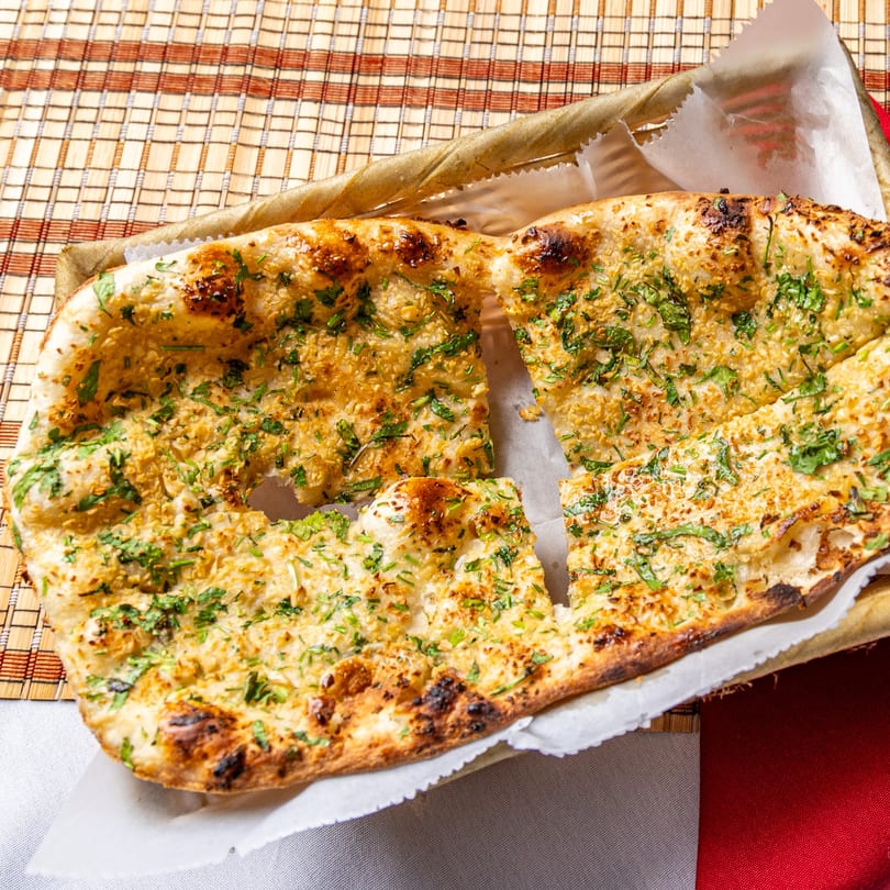 Golden-brown garlic naan bread, topped with green herbs, served in a basket on a woven placemat.