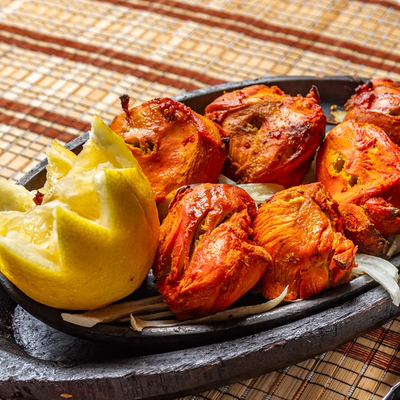 Tandoori chicken pieces with lemon and onions on a dark platter, against a woven placemat.
