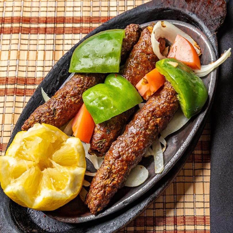 Indian seekh kebabs with green bell peppers, onions, tomatoes, and a squeezed lemon on a dark platter.