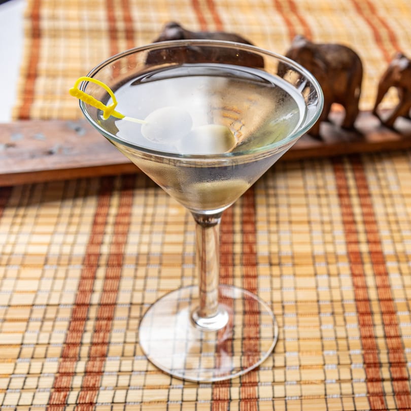 Martini with olives and a yellow sword pick on a woven placemat with elephant figurines in the background.