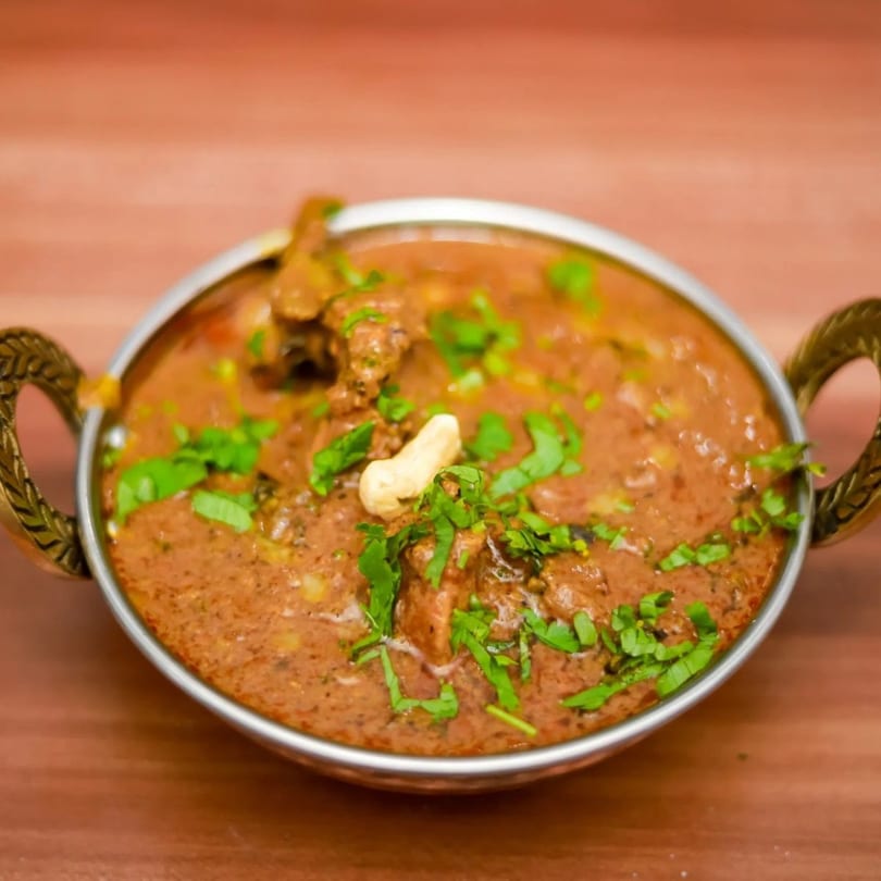 Indian curry with meat, cilantro, and a cashew in a metal bowl on a wooden table.